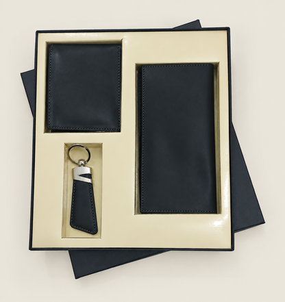 A three-piece men's leather gift set in a cream-colored presentation box, including a slim black leather wallet, a matching long mobile wallet, and a black leather keychain with a silver accent.